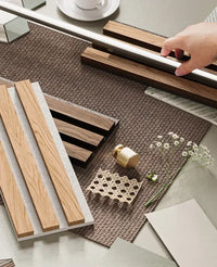 Hand arranging wooden beams and decor pieces on a textured surface with small white flowers and a cup in the background.