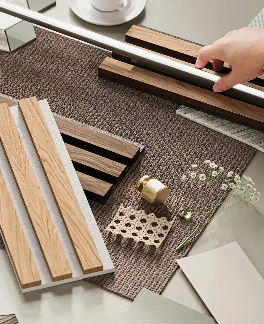 Hand arranging wooden beams and decor pieces on a textured surface with small white flowers and a cup in the background.